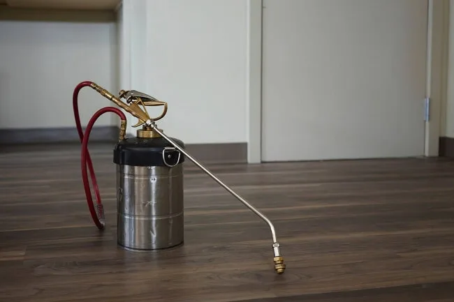 Pest Management Spray for Spider Removal A metal pesticide sprayer with a long nozzle and red hose is placed on a wooden floor in a room with beige walls and a closed door. The sprayer has a brass pump handle on top.