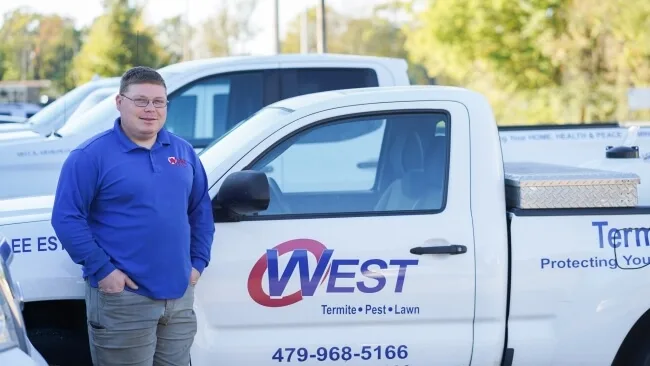Silverfish removal expert standing by truck A man in a blue shirt stands smiling next to a white pickup truck with West Termite Pest Lawn written on the door. The truck is parked outdoors on a sunny day with trees in the background.