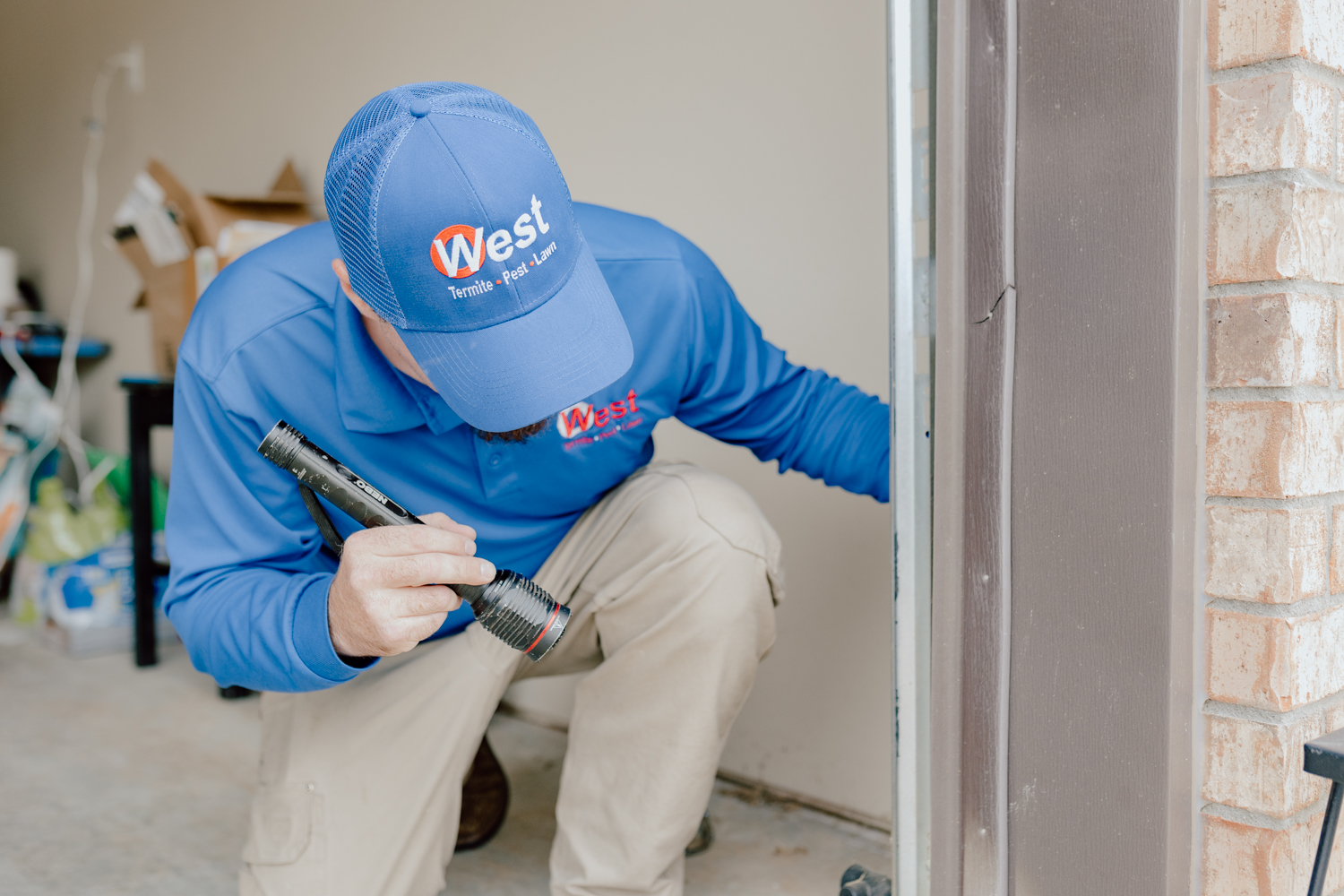 A person in a blue shirt and cap labeled West examines a door frame with a flashlight. Theyre kneeling in a garage area with a brick wall nearby and various items in the background.