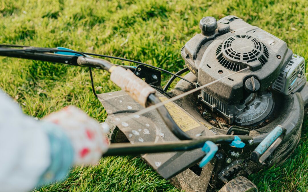 Close-up of a person starting a gas-powered lawn mower on green grass, with their hands pulling the starter cord and the engine visible.