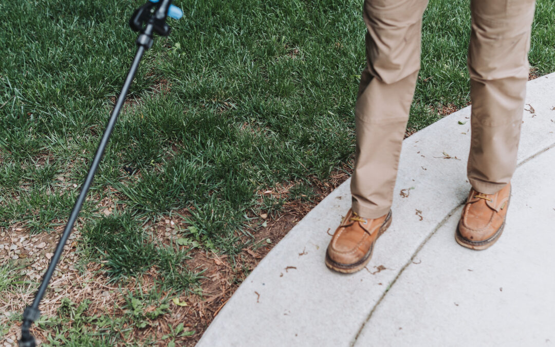A person wearing tan pants and brown boots stands on the edge of a sidewalk next to green grass, holding a sprayer wand pointed at the ground.
