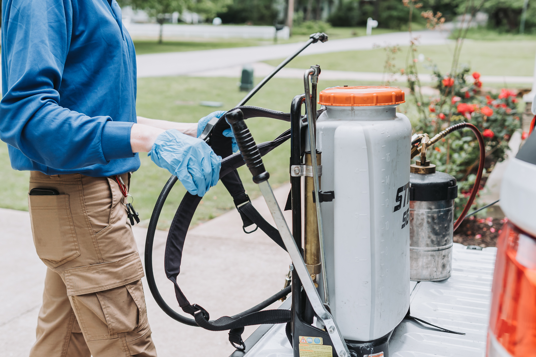 A person wearing blue gloves and a blue shirt is handling a large backpack sprayer and a metal canister on the back of a truck in a residential area, preparing for termite inspections or outdoor pest control to protect real estate.