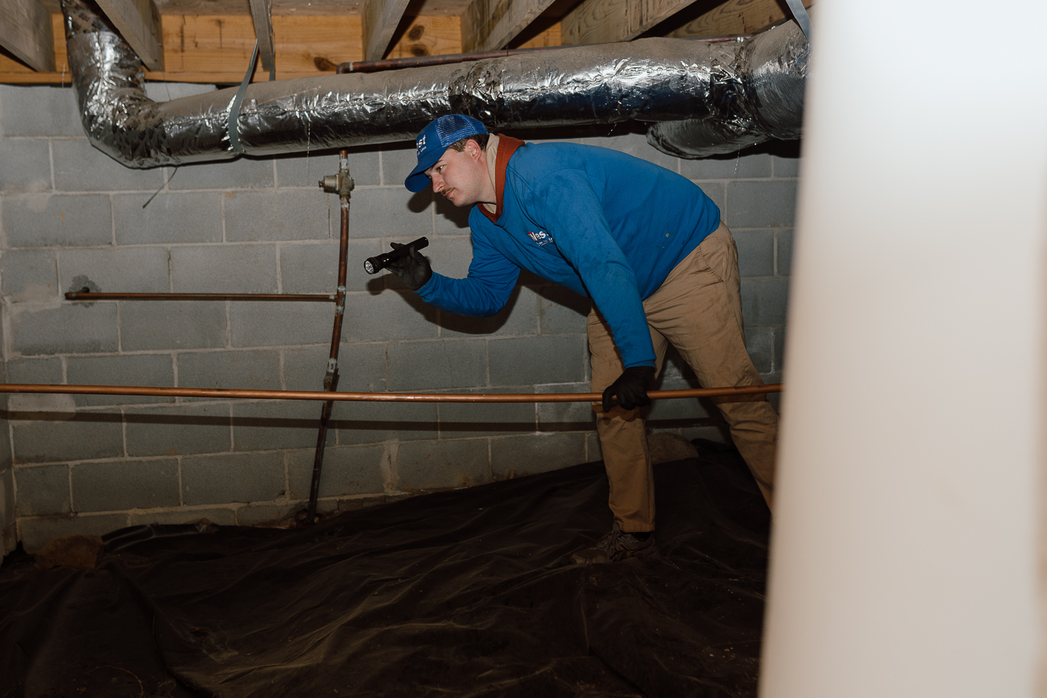 Post #06 A person wearing a blue jacket, khaki pants, gloves, and a blue cap inspects a crawl space with a flashlight during a termite inspection, surrounded by pipes and cinder block walls.
