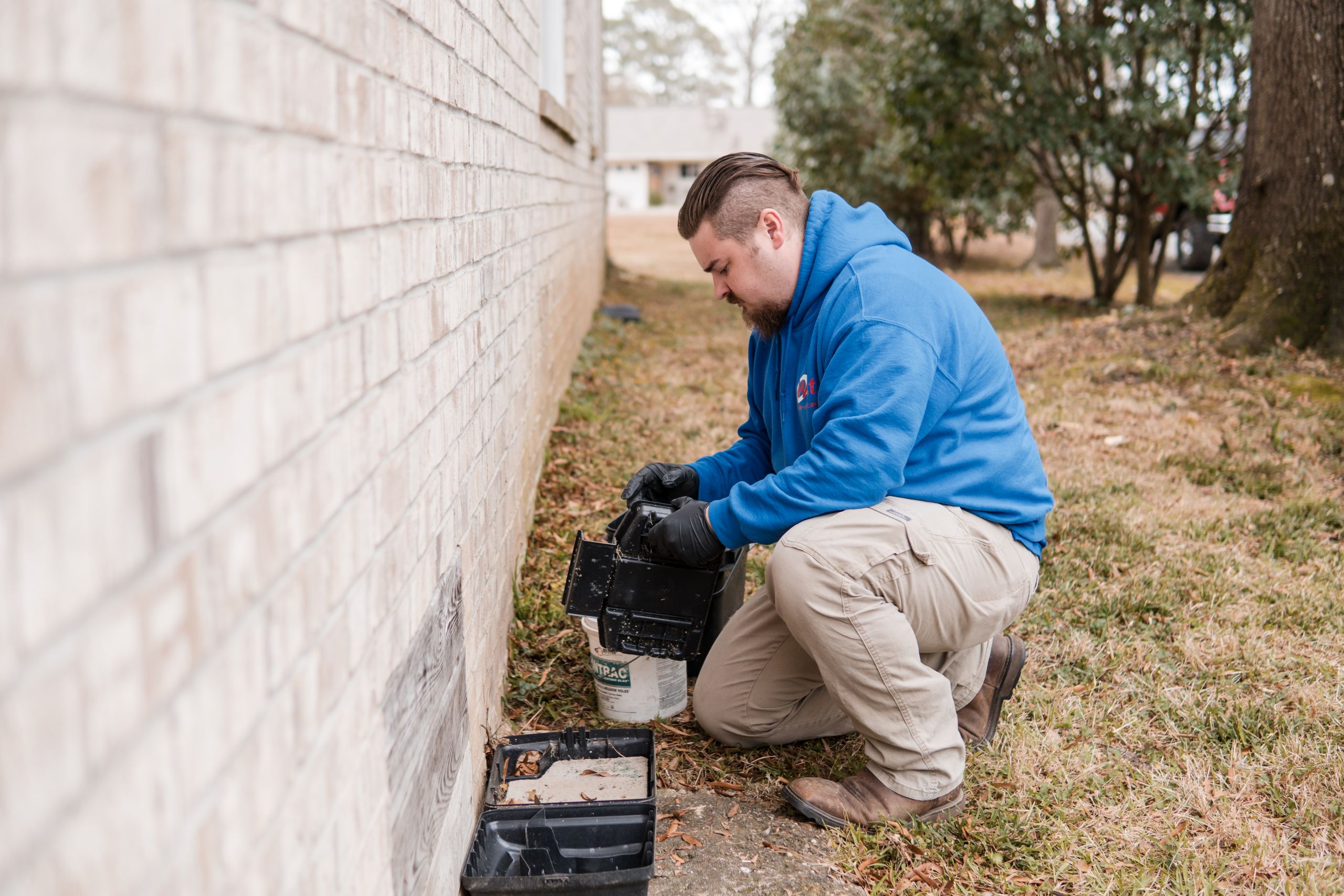 A man in a blue hoodie and khaki pants kneels on the grass next to a brick building, checking a black bait station used for termite treatment near the foundation. Trees and houses can be seen in the background.