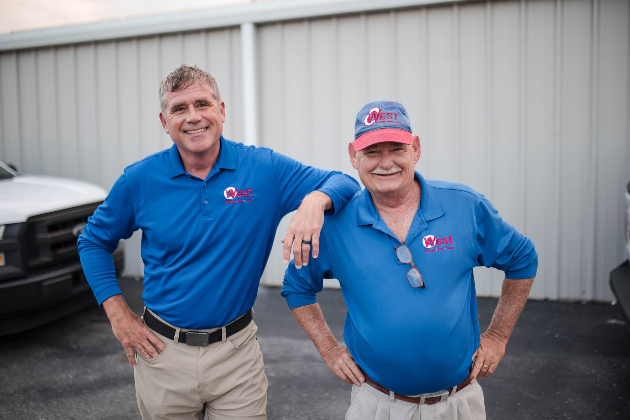 Two men in blue West logo shirts and beige pants stand smiling outside a metal building, ready to provide termite protection. One wears a cap and sunglasses, and both have their hands on their hips. Vehicles are partially visible in the background.