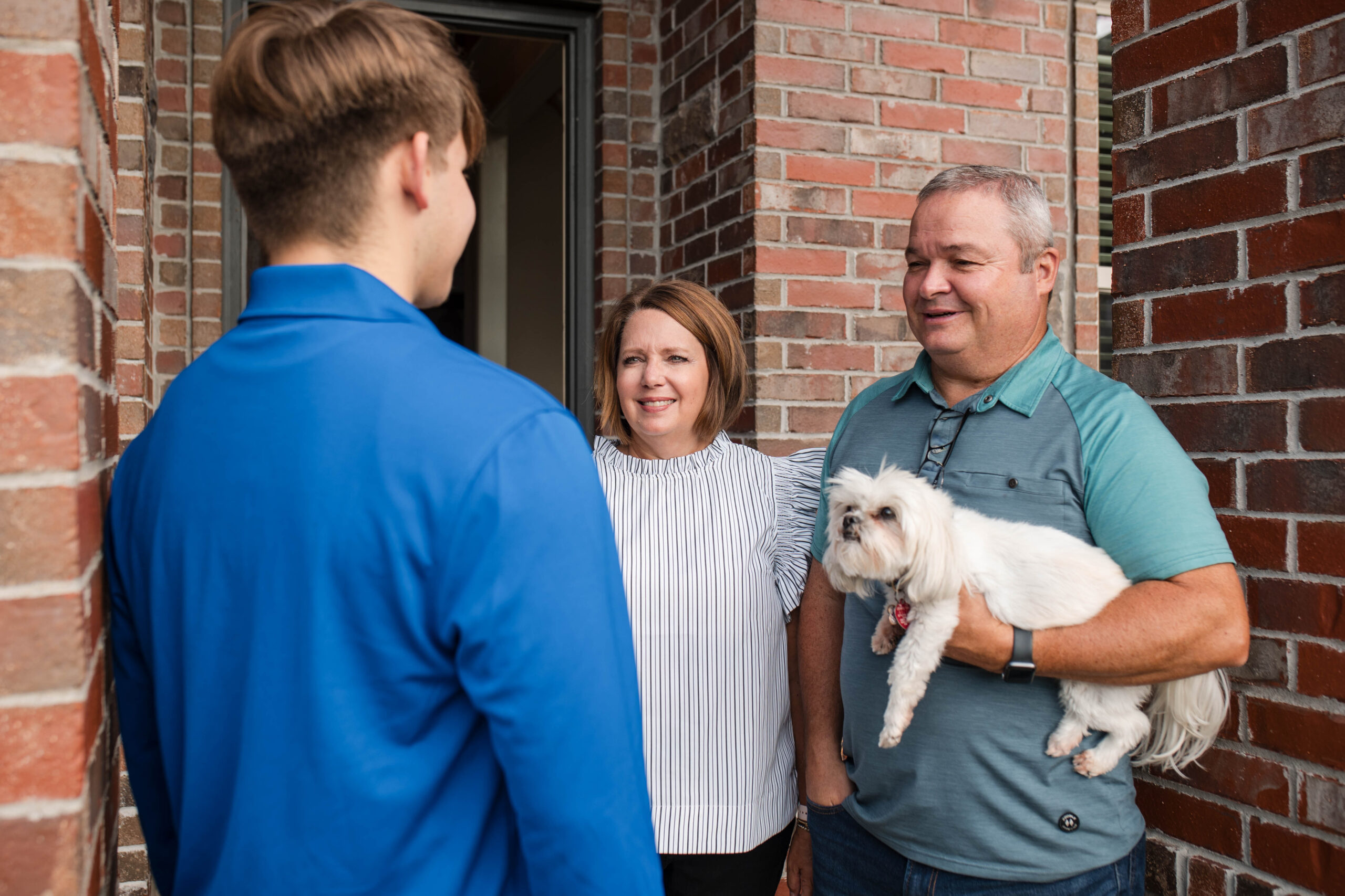 A young man in a blue shirt stands at a doorway, speaking to a smiling middle-aged couple holding a small white dog, likely discussing their termite problem as they greet each other outside a brick house.