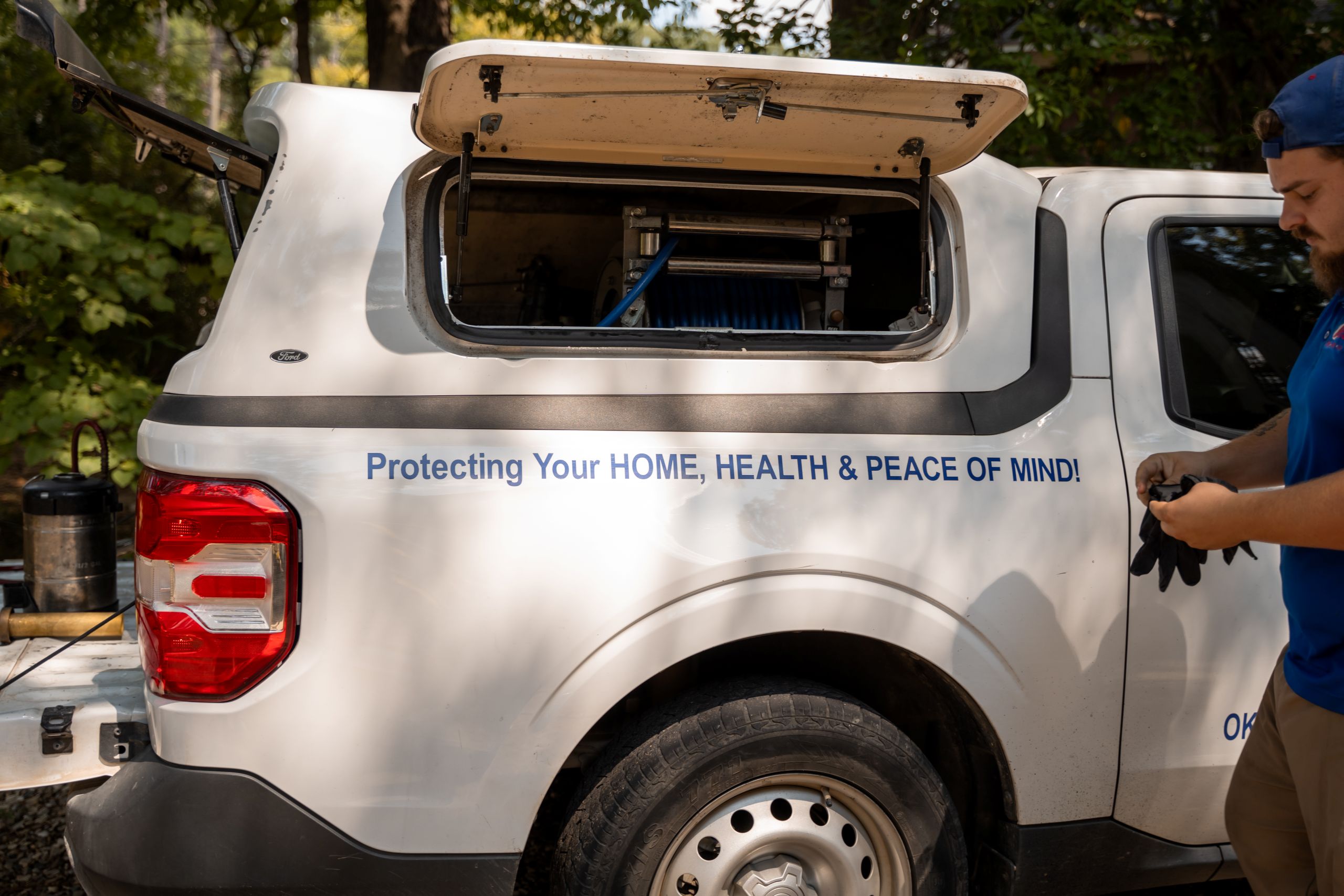 A white service truck with an open side compartment displays the text: Protecting Your HOME, HEALTH & PEACE OF MIND from pests! A man in a blue shirt and cap stands next to the truck, putting on black gloves. Trees are visible in the background.