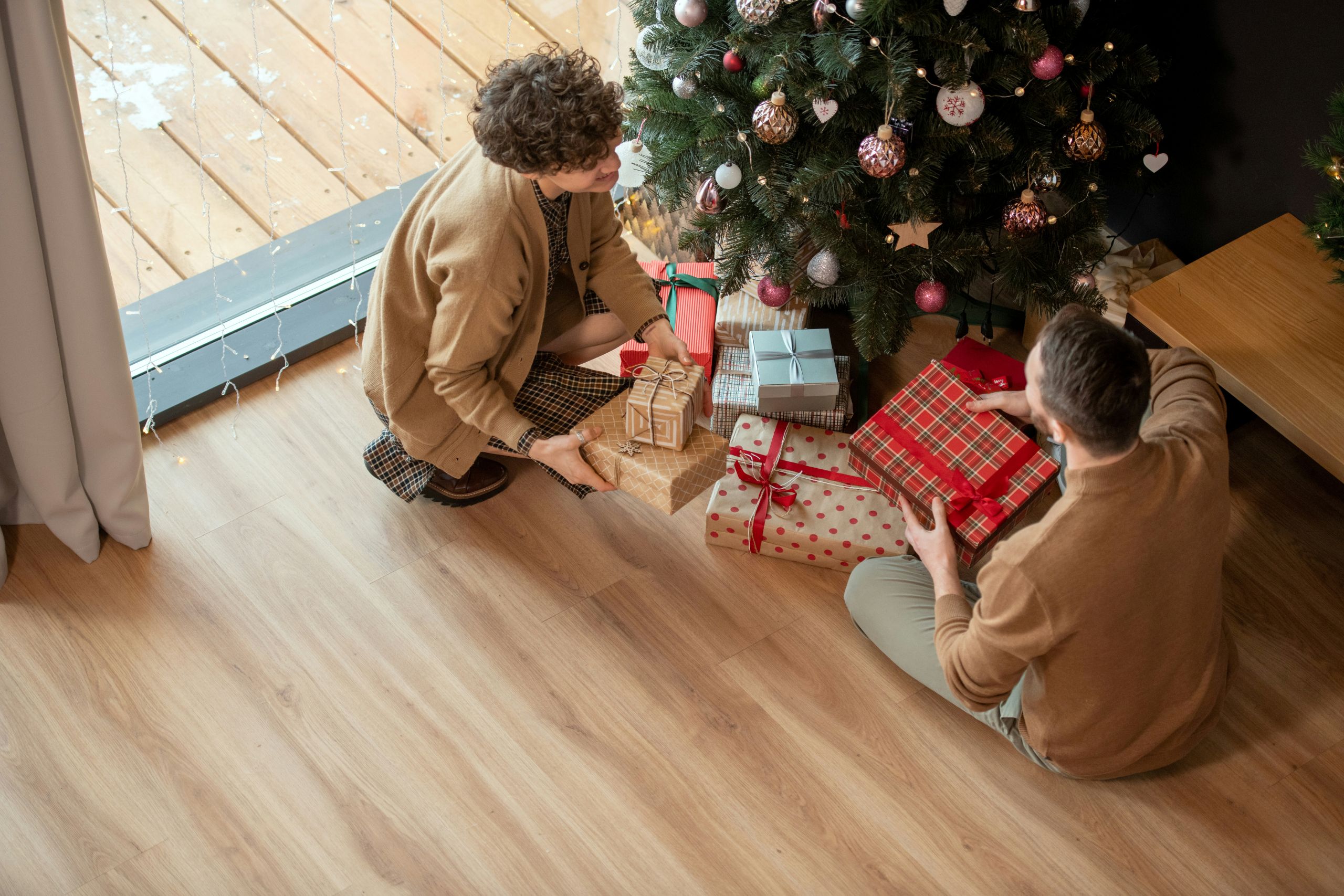 Two people sit on a wooden floor near a decorated Christmas tree, arranging wrapped gifts with various patterns under the tree, making sure everything is perfect and free from pests as light streams in from a nearby window.