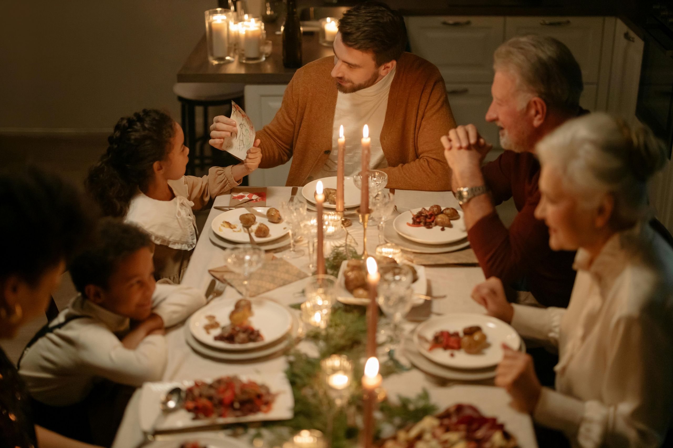A family gathers around a candlelit dinner table, sharing a meal and smiling. As adults hand something to children and others chat, the warm, festive atmosphere keeps thoughts of winter pests far from their cozy gathering.