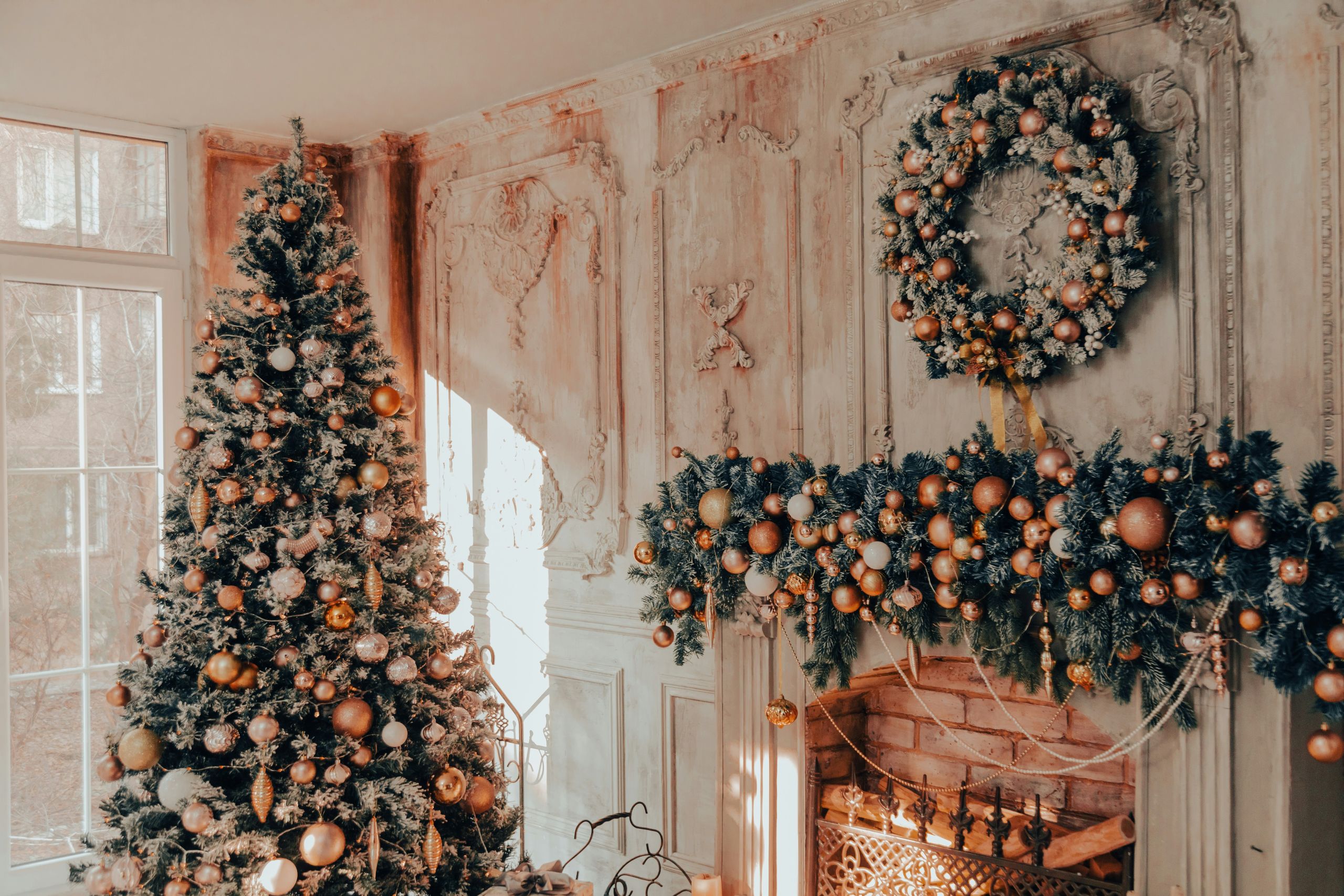 A decorated Christmas tree stands by a large window beside a fireplace, adorned with a festive garland and wreath. Elegant, vintage-style white walls and soft natural light create a cozy holiday scene—perfect tips to help Keep Your Home Pest-Free this season.
