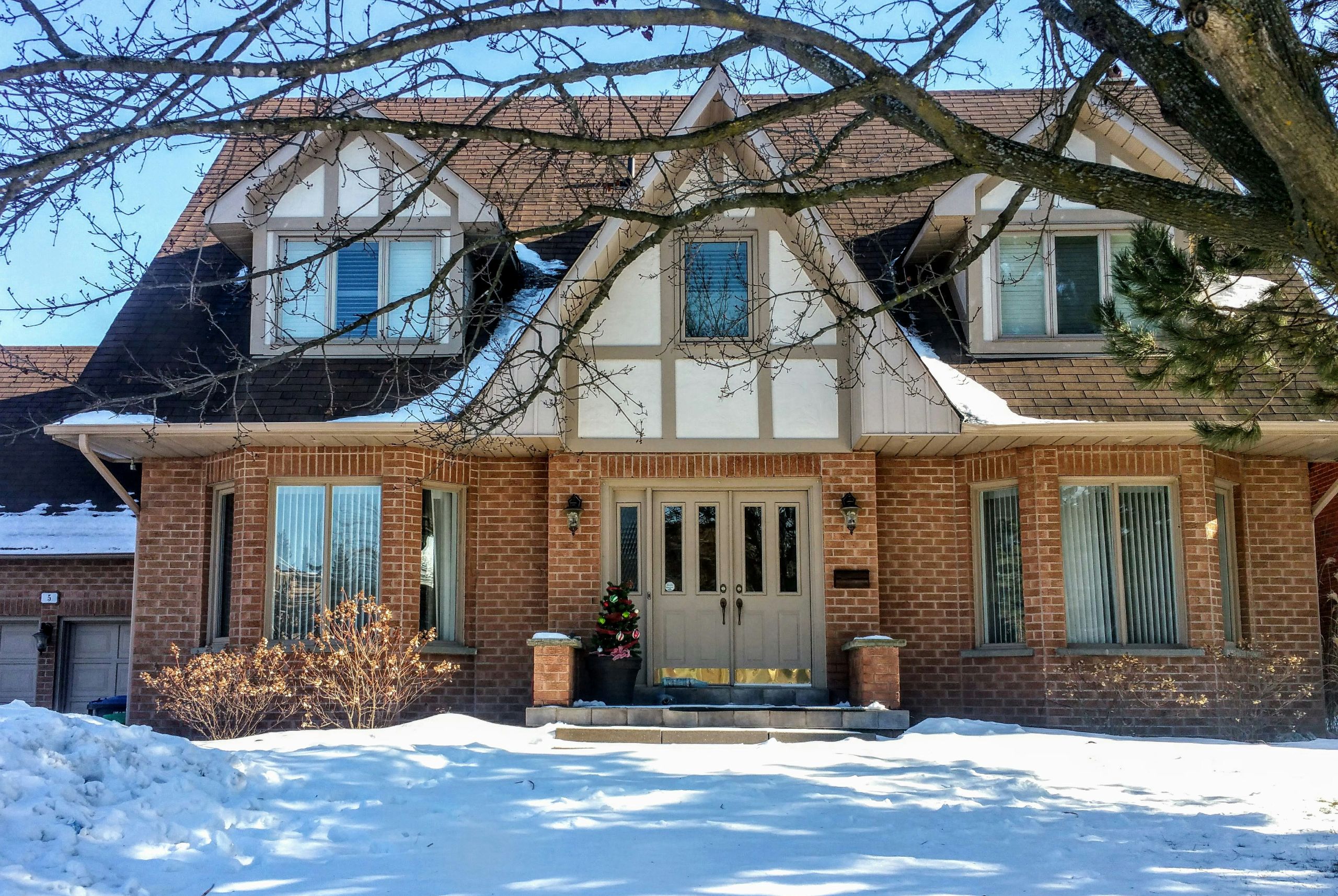 A two-story brick house with snow covering the front yard and steps. Bare tree branches frame the scene, and a small decorated Christmas tree sits by the front door under a clear blue sky—a serene winter termite haven.