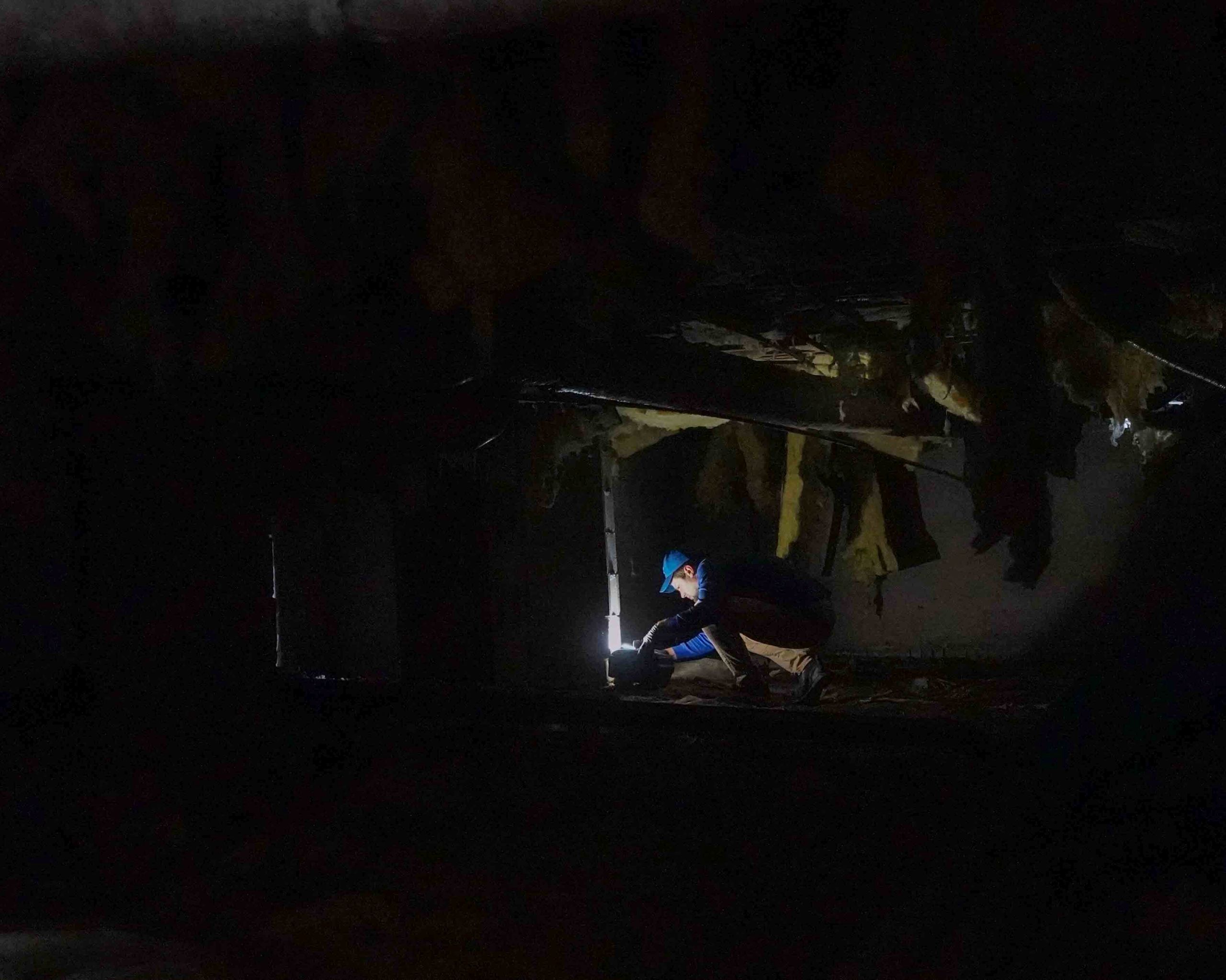 A person in a blue hat crouches in a dark, cluttered basement or crawl space, illuminated by a small flashlight, searching for signs of pests indoors with insulation hanging from the ceiling above.