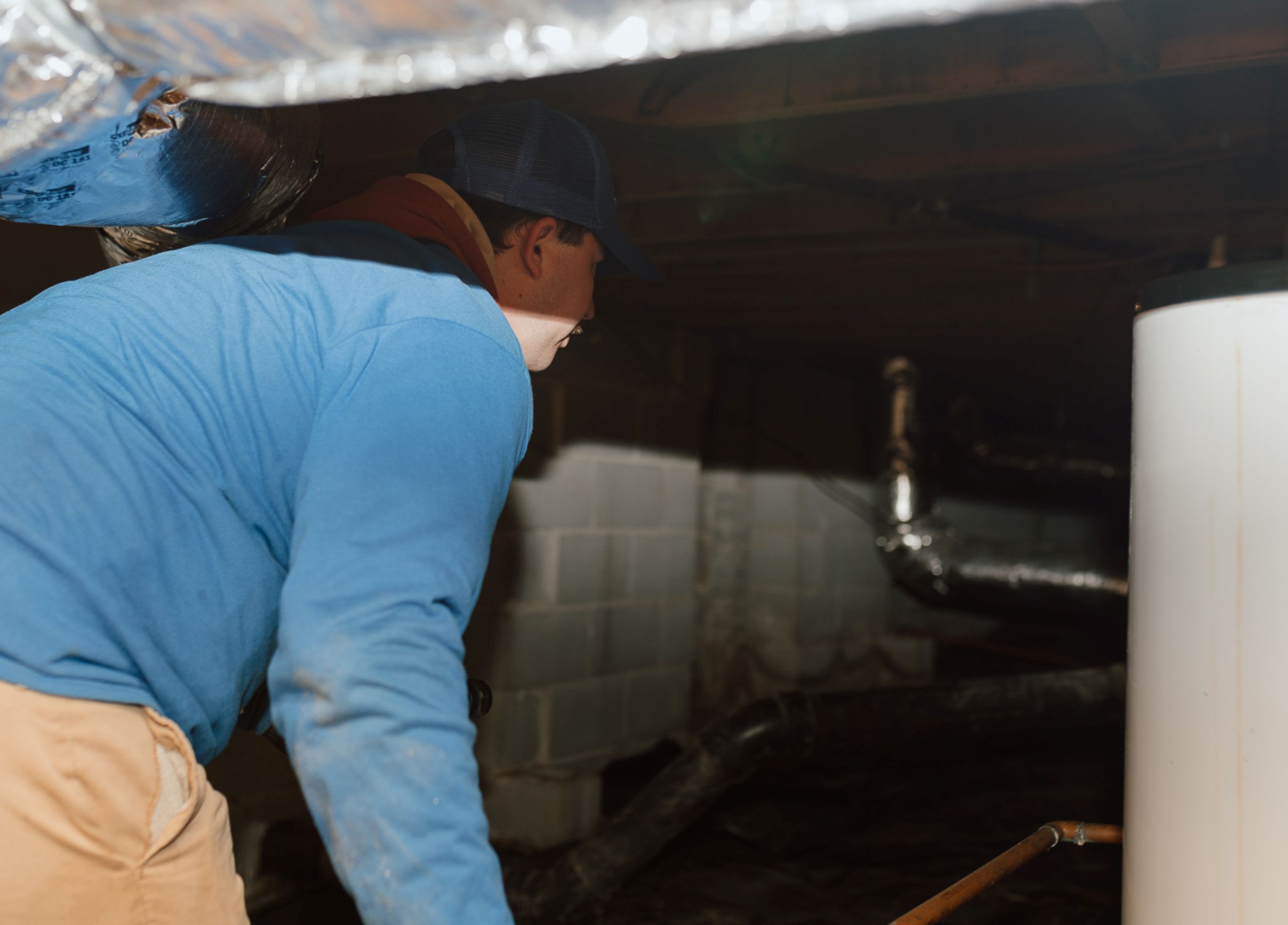 Why Crawl Spaces Are Vulnerable in Winter A person wearing a blue shirt and cap works in a dimly lit crawl space with exposed pipes, a water heater, and concrete block walls—an area often vulnerable to winter pests.