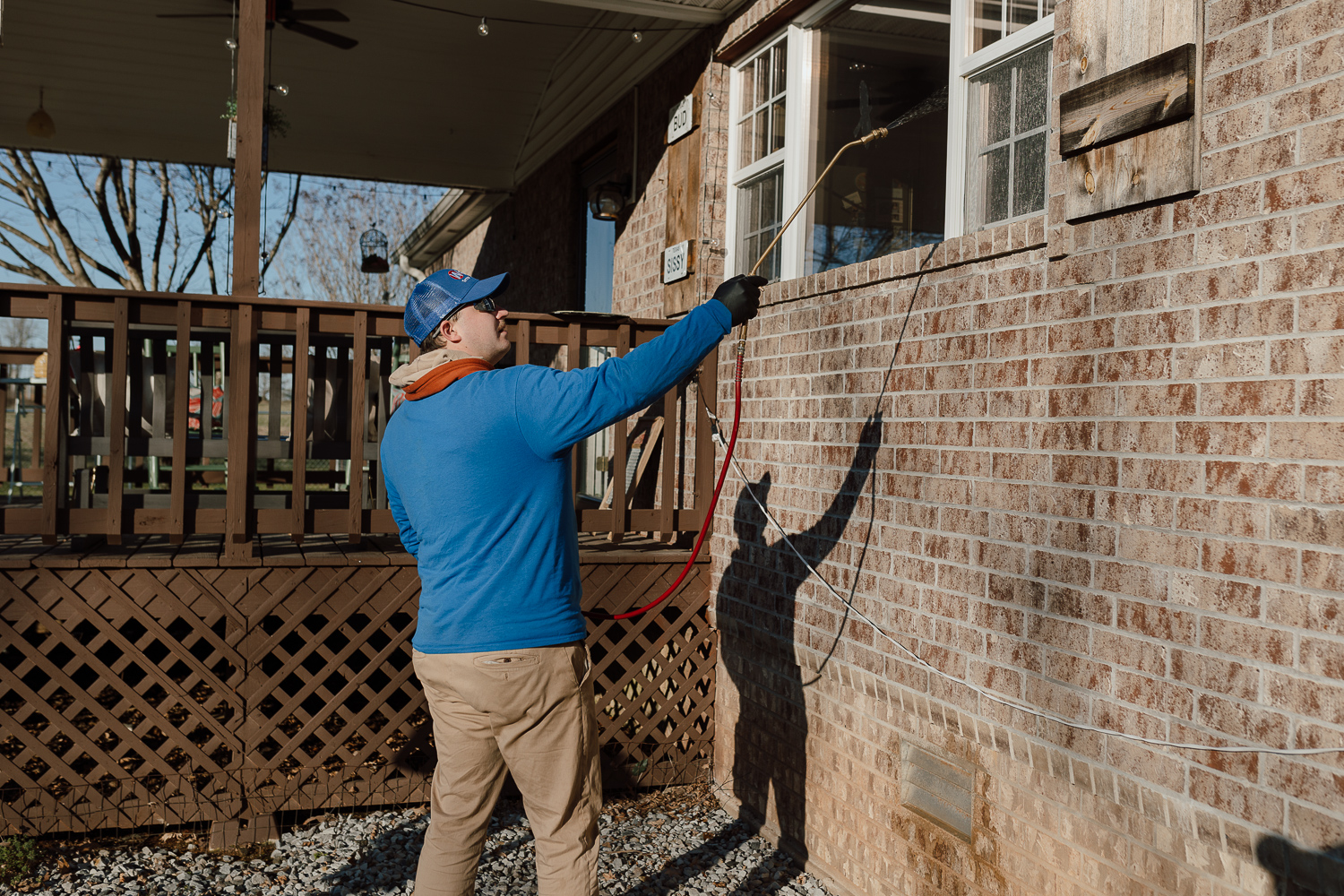 Pestproofing Entry Points Before Spring A person wearing a blue cap, blue shirt, and gloves sprays liquid onto the brick exterior of a house near a window, possibly pestproofing entry points before spring or performing cleaning.