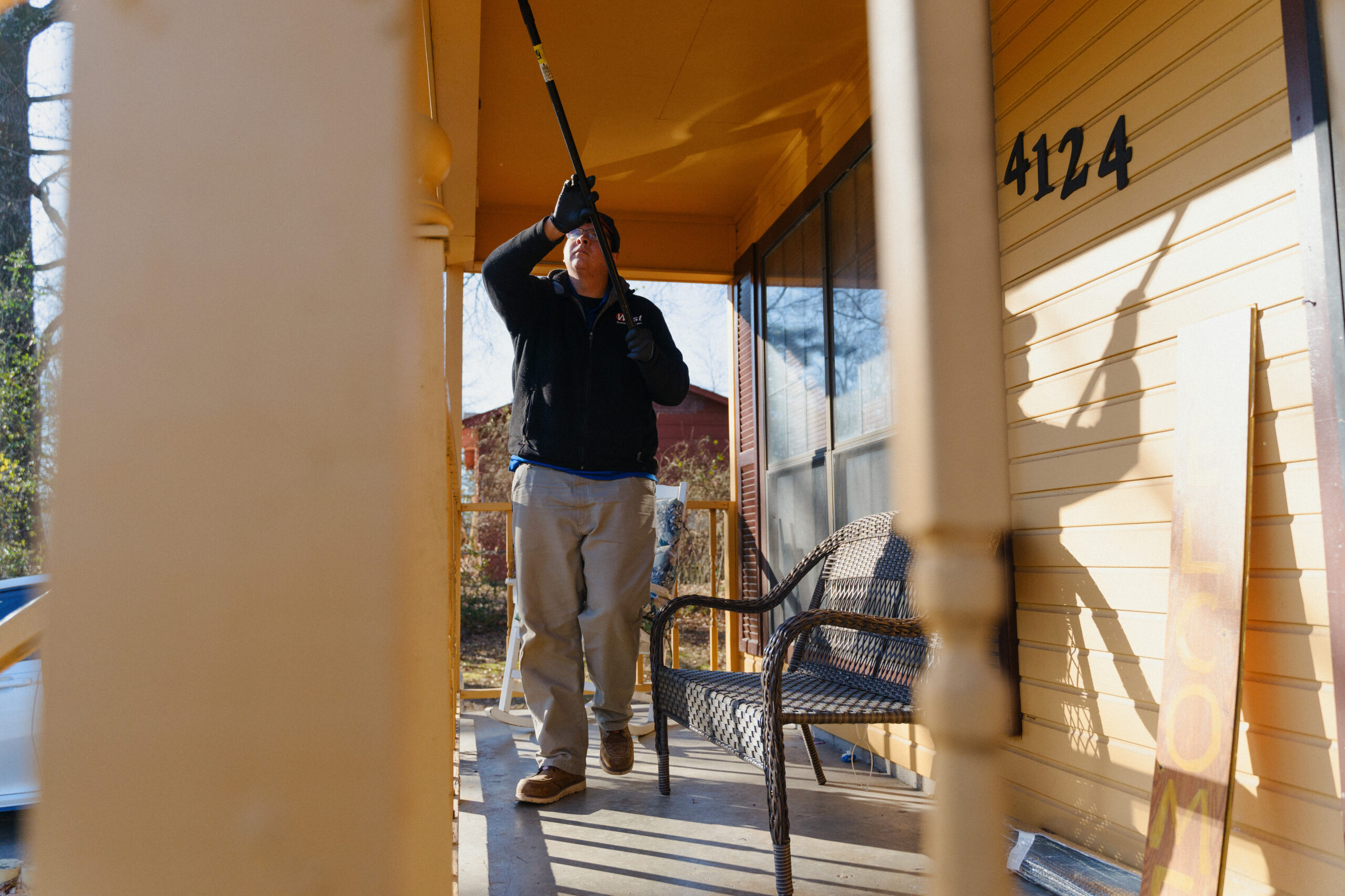 A person wearing gloves and a jacket uses a long-handled tool to clean the ceiling of a yellow house’s porch, near a wicker chair and a house number sign reading 4124, preparing your home for early spring pests.