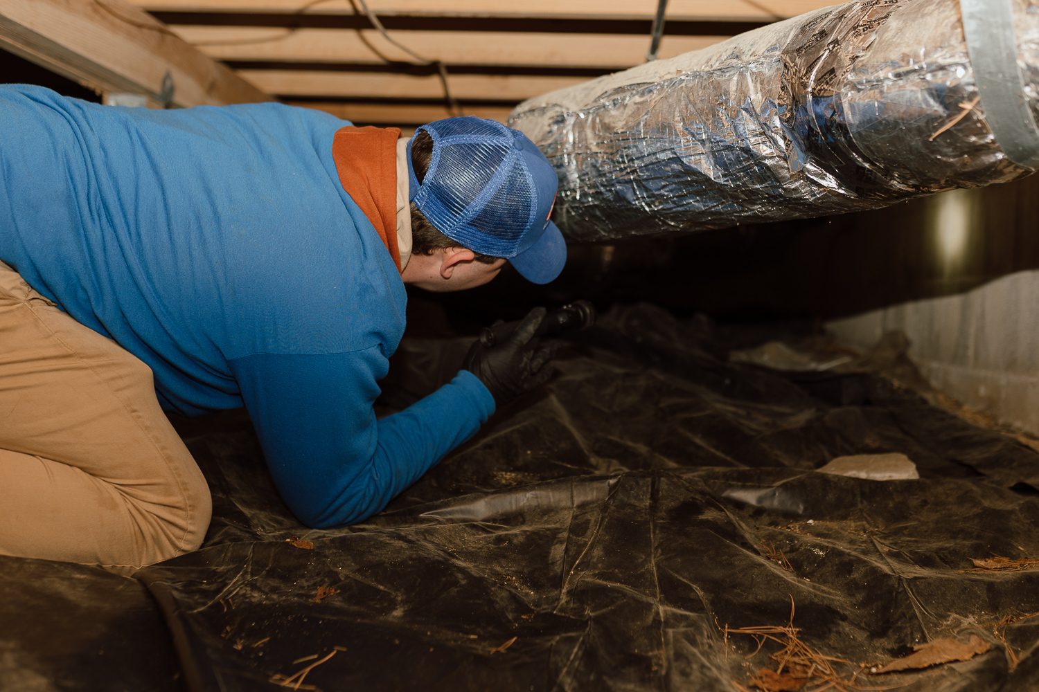 Preventing Cockroach, Silverfish, and Termite Problems A person in a blue shirt, brown pants, and blue cap is crawling in a crawl space under a house, possibly checking for termite problems or working near an insulated air duct. The ground is covered with black plastic sheeting.