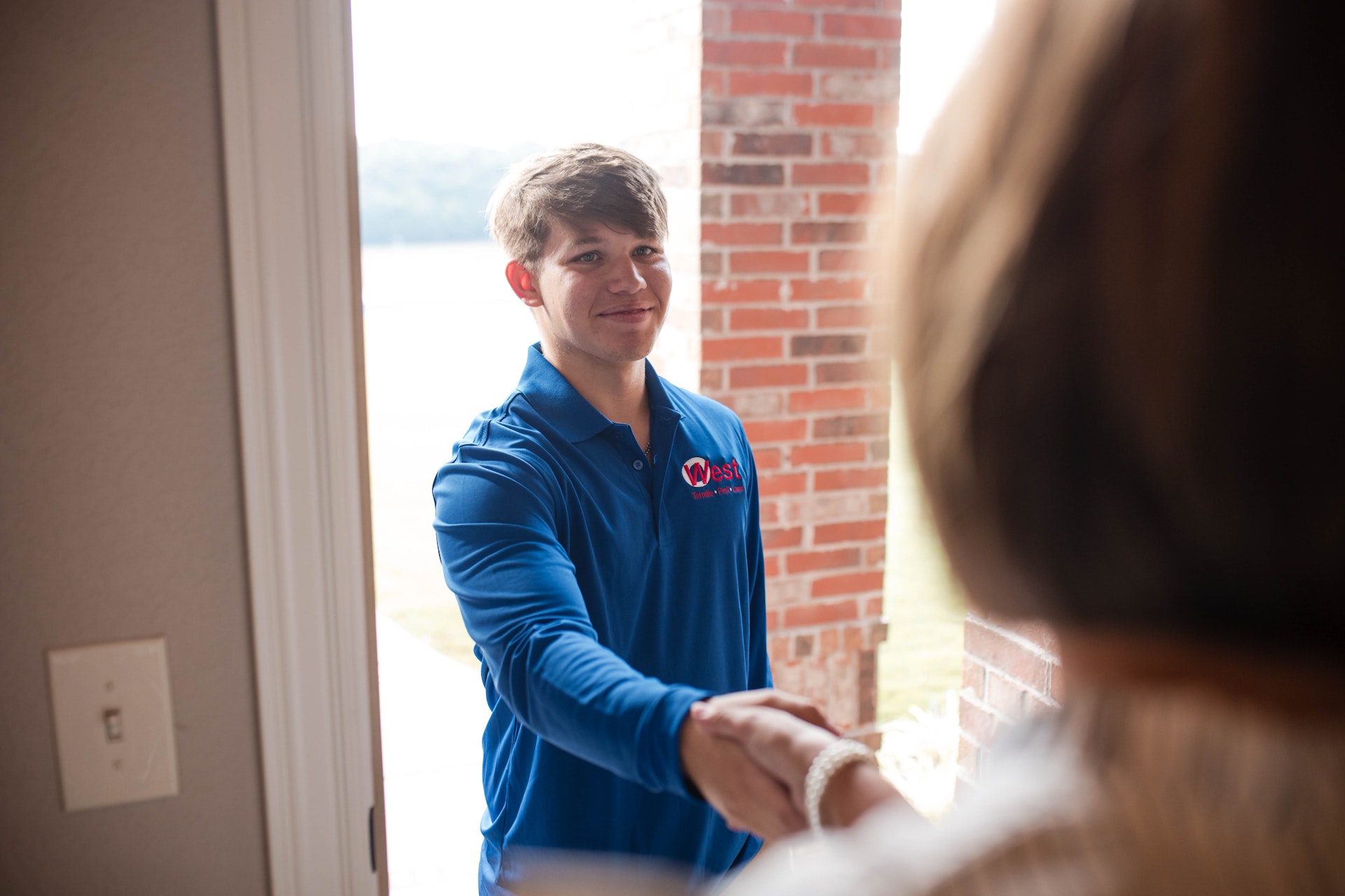 A man in a blue polo shirt stands at a doorway, smiling and shaking hands with a person whose back is to the camera, with a brick wall and outdoor scenery visible in the background.