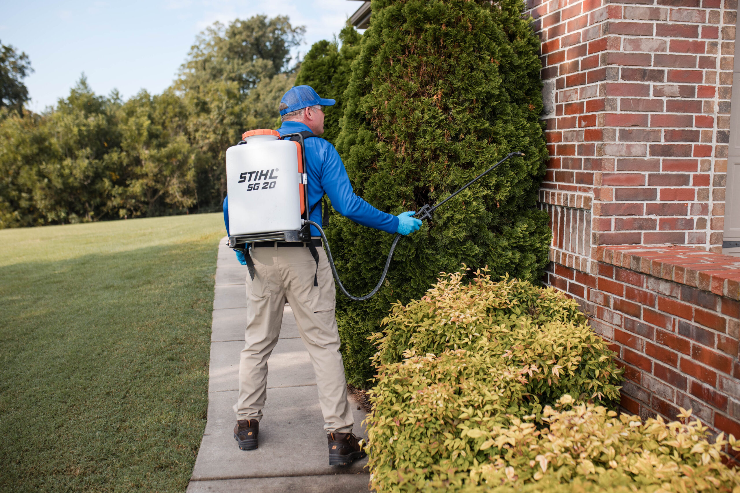 A person wearing a blue cap and shirt uses a STIHL SG 20 backpack sprayer to treat bushes next to a brick house on a sunny day.