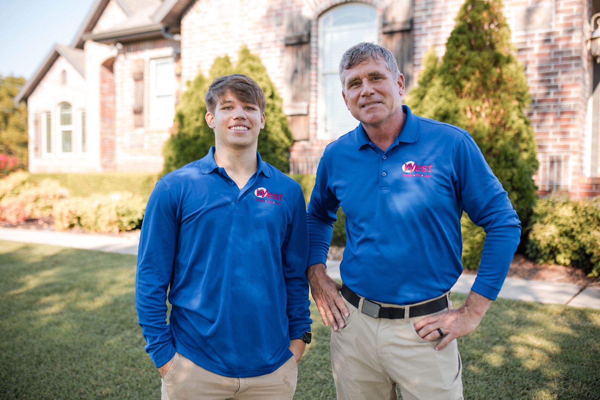 Two men in matching blue shirts with a red and white logo stand outside in front of a brick house and green bushes, posing and smiling for the camera.