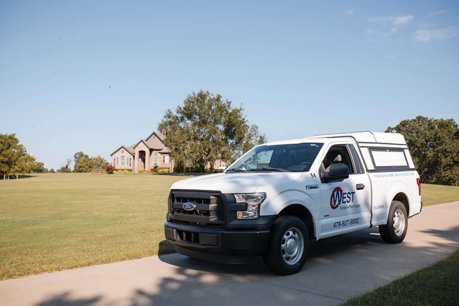 A white West Plumbing service truck parked on a driveway in front of a large house with a well-kept lawn and trees under a clear blue sky.
