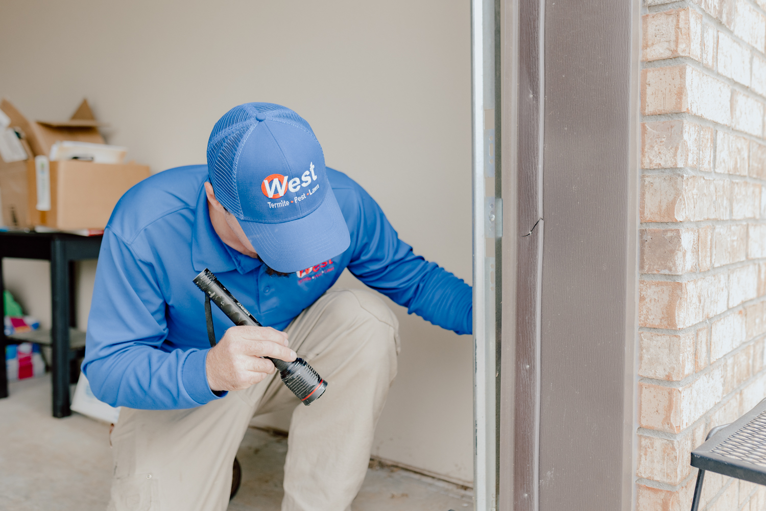 A person wearing a blue West cap and shirt uses a flashlight to inspect the bottom corner of a garage door near a brick wall.