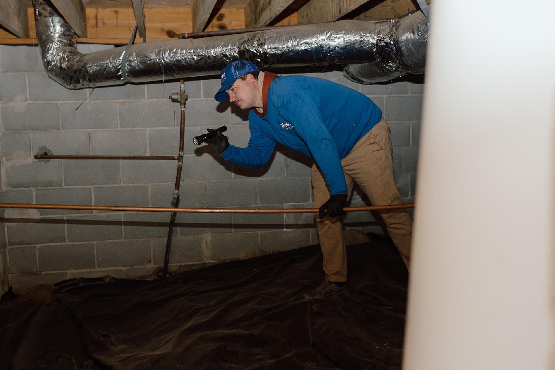 A worker in a blue jacket and hat inspects pipes with a flashlight in a dimly lit basement with exposed brick walls and ductwork.