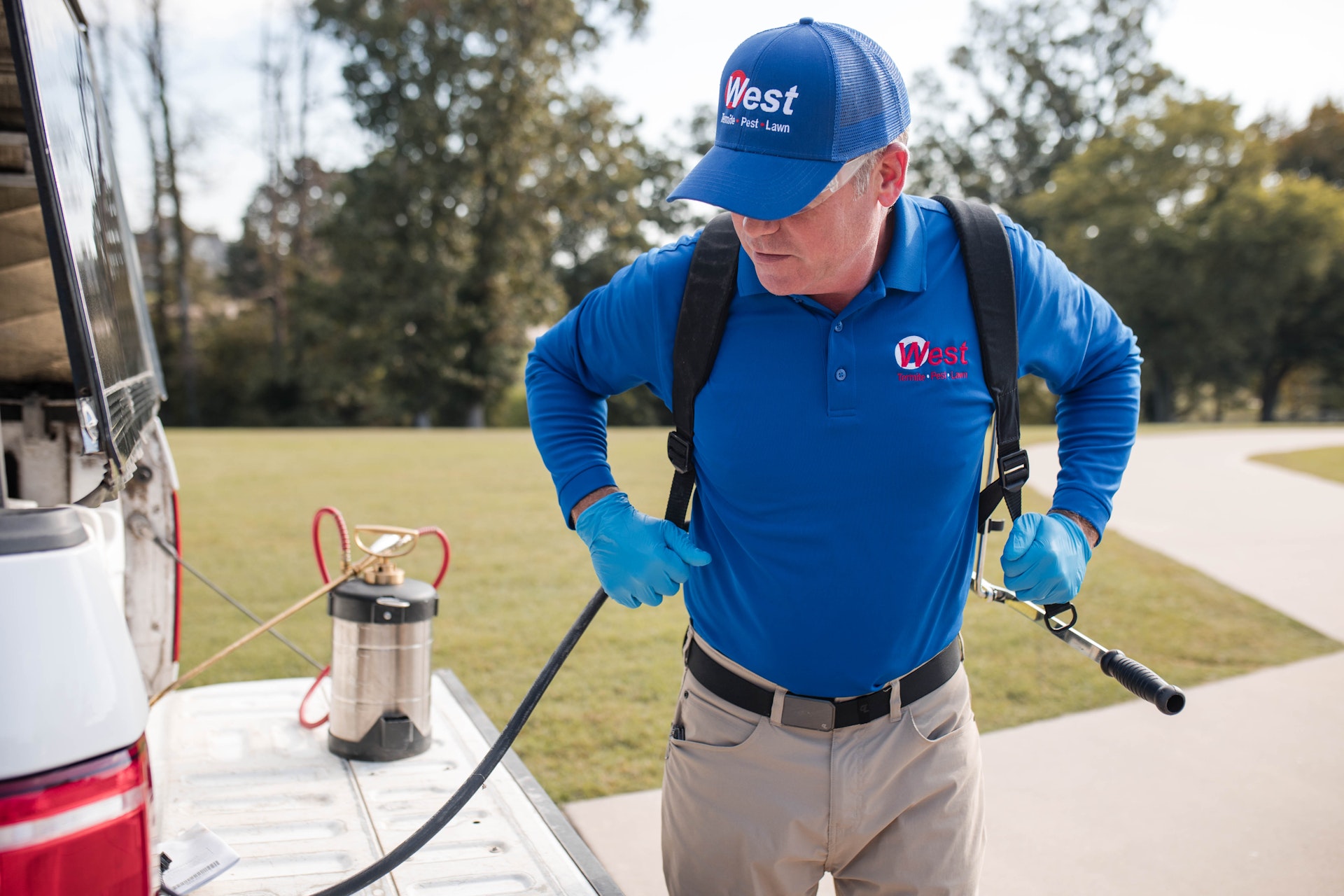 A pest control worker in a blue uniform and gloves puts on a backpack sprayer next to a truck, preparing equipment for use outdoors on a sunny day.