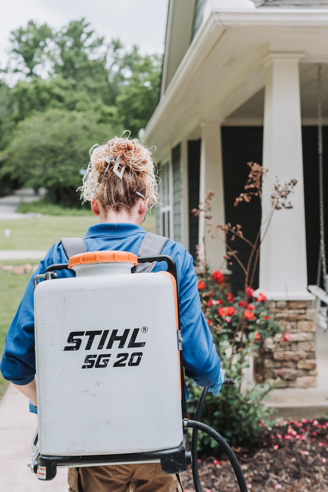 Post #03_2 A person with curly hair, wearing a blue shirt, carries a STIHL SG 20 backpack sprayer while walking on a path toward a house with a porch and a garden of red flowers.