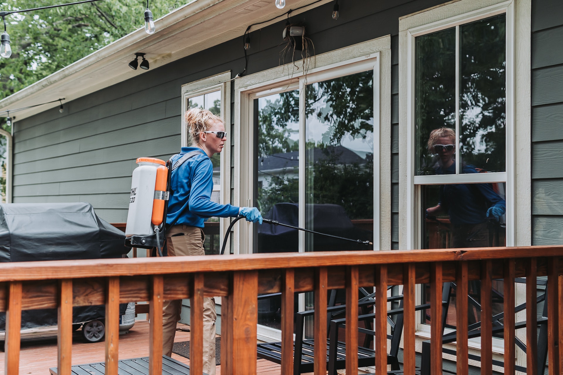 A person wearing gloves and safety glasses sprays a house exterior with pest control equipment on a deck, near large windows and a barbecue grill.