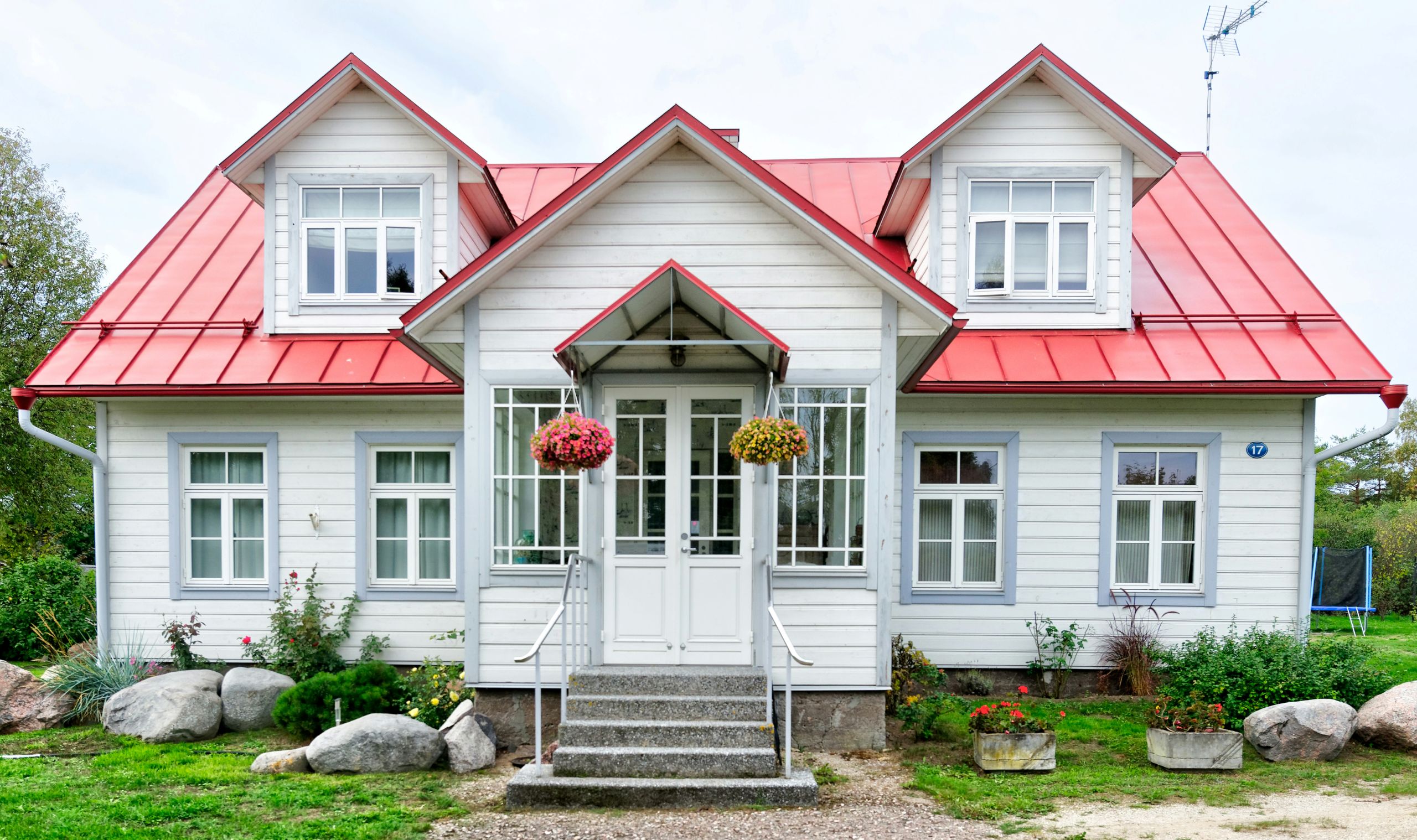 A white house with a red metal roof, two dormer windows, a small front porch with glass panels, hanging flower baskets, stone landscaping, and a grassy yard free from signs of pest damage.