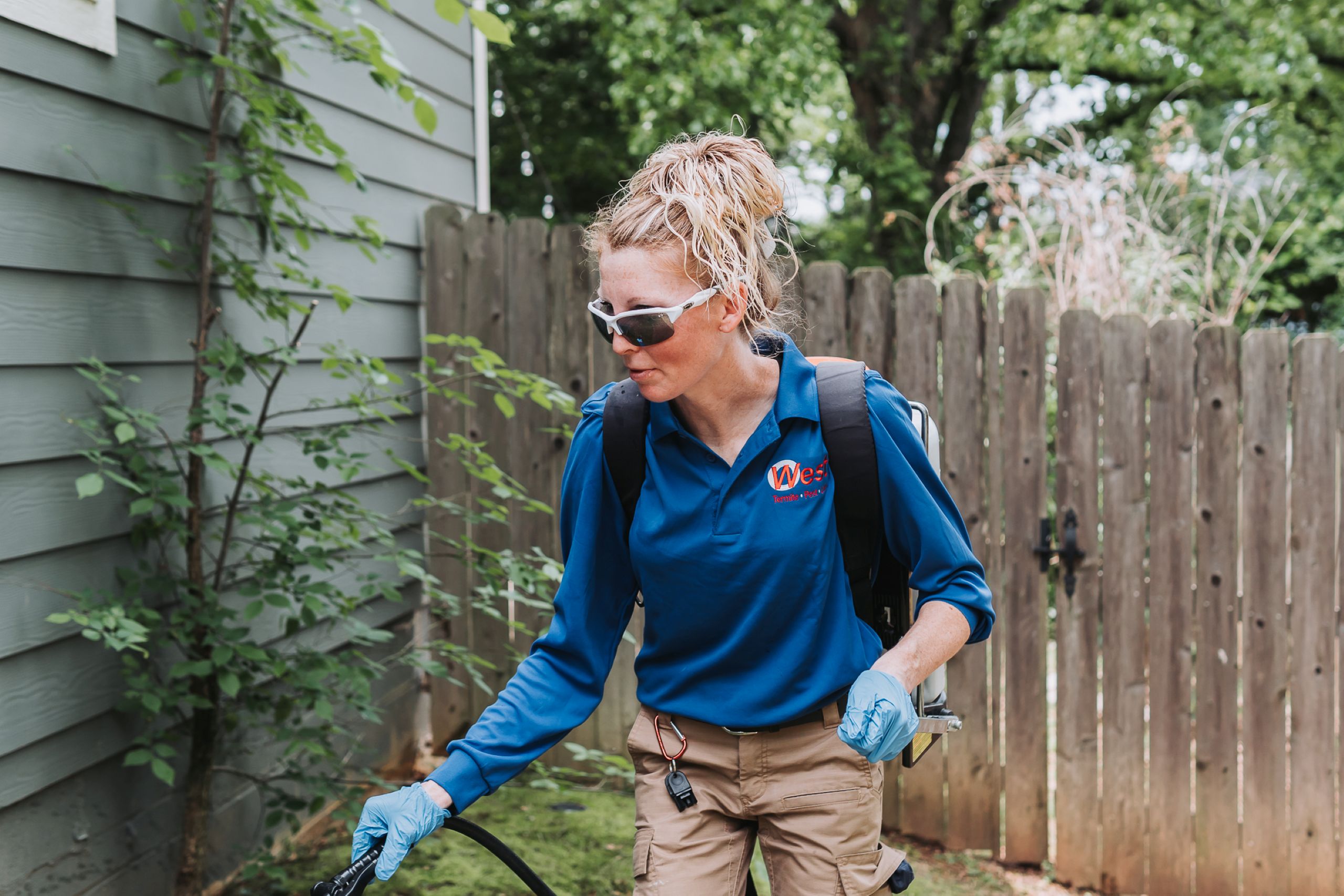 A woman in sunglasses, gloves, and a blue uniform uses spring pest control equipment outdoors, spraying near a house and wooden fence on a green lawn.