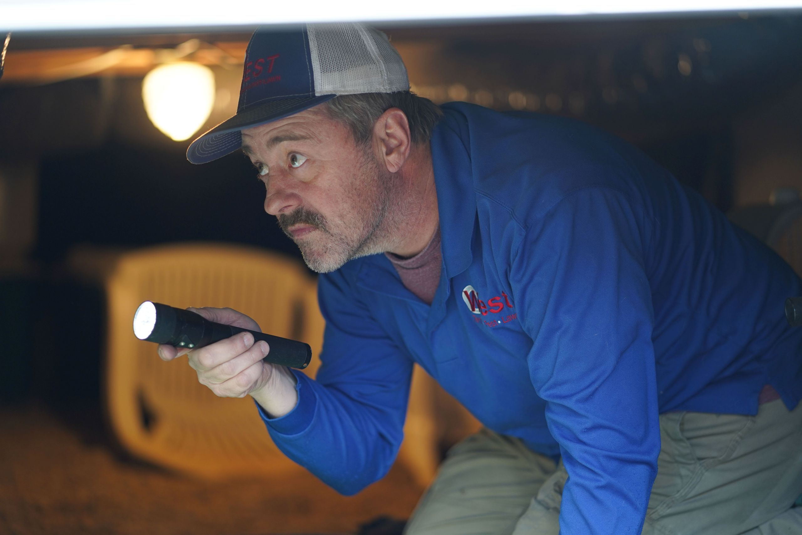 A man wearing a blue shirt and cap crouches under a structure, holding a flashlight and carefully inspecting for pests—an essential step for spring cleaning. A chair and light bulb are visible in the background.
