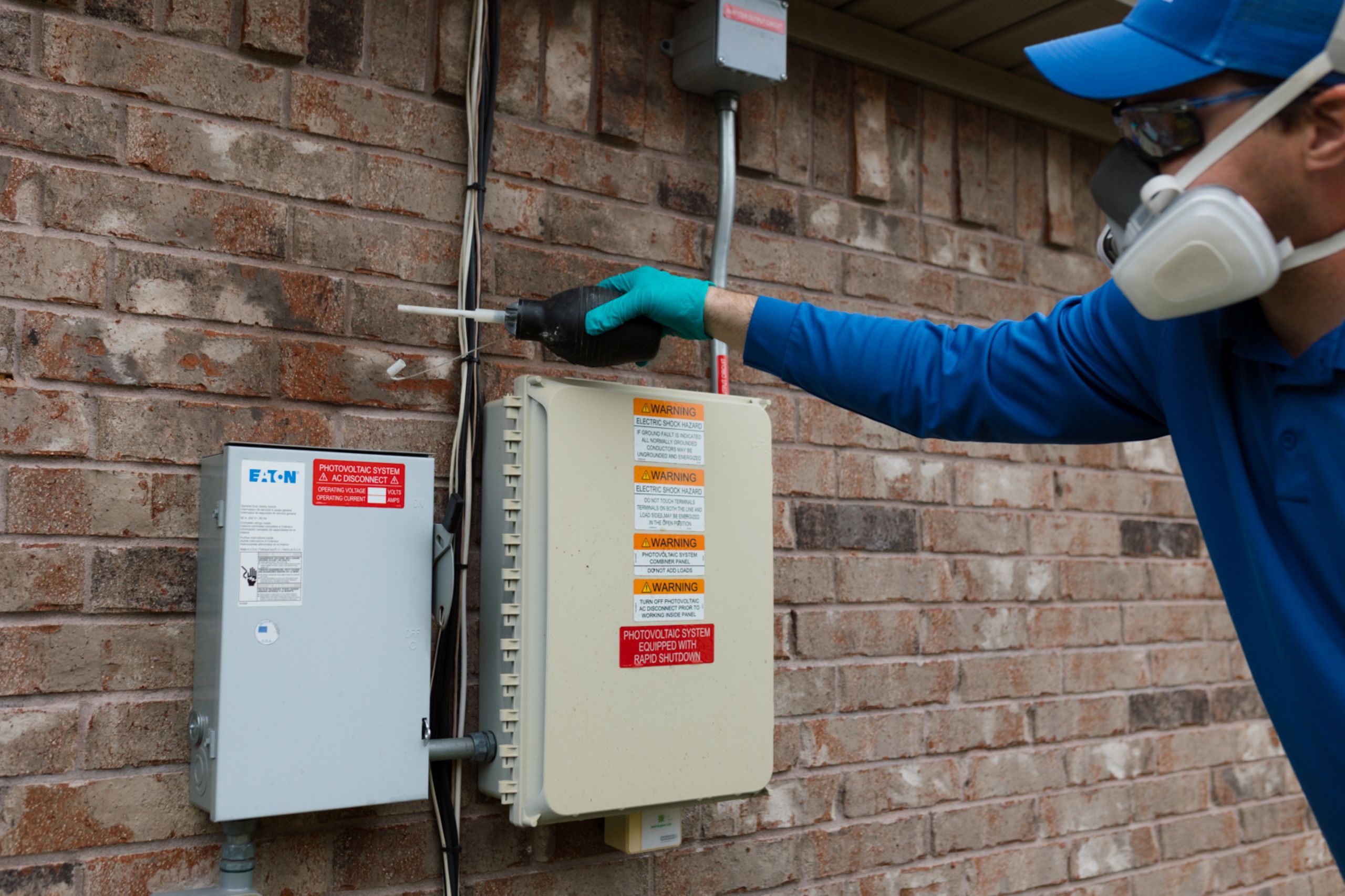 A person wearing safety gear uses a tool to inspect electrical or utility boxes mounted on a brick wall outside a building for preventative pest control.