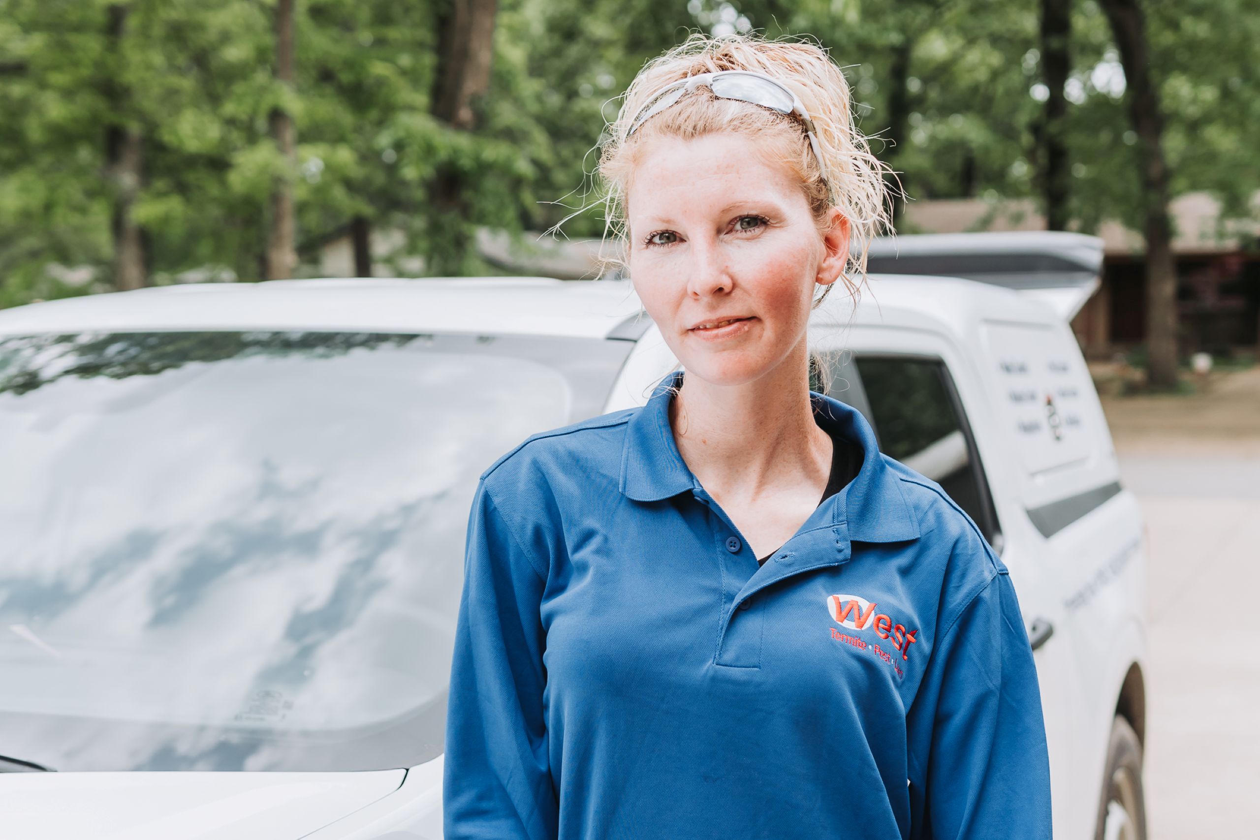 A woman with blonde hair pulled back, wearing a blue polo shirt, stands in front of a white vehicle outdoors. The vehicle has writing on its side, possibly related to soil conditions, with trees and a house visible in the background.