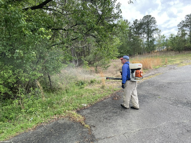 Post #04 (1) A person using a backpack sprayer applies chemicals to vegetation near a paved area, surrounded by trees and grass under a cloudy sky.