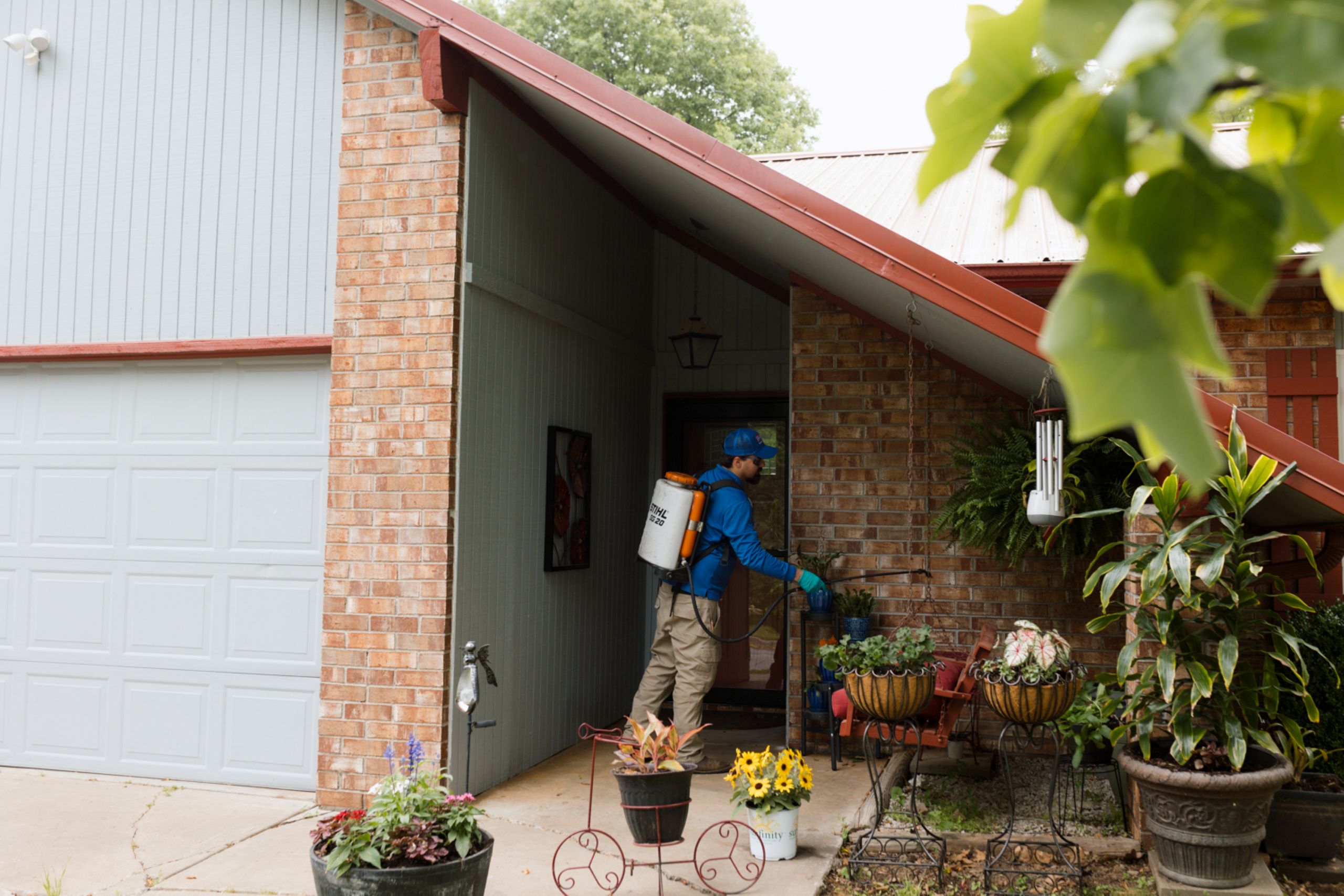 Why Annual Termite Inspections Are Worth It A person in blue spraying plants with a backpack sprayer on the porch of a brick house surrounded by potted flowers and greenery.
