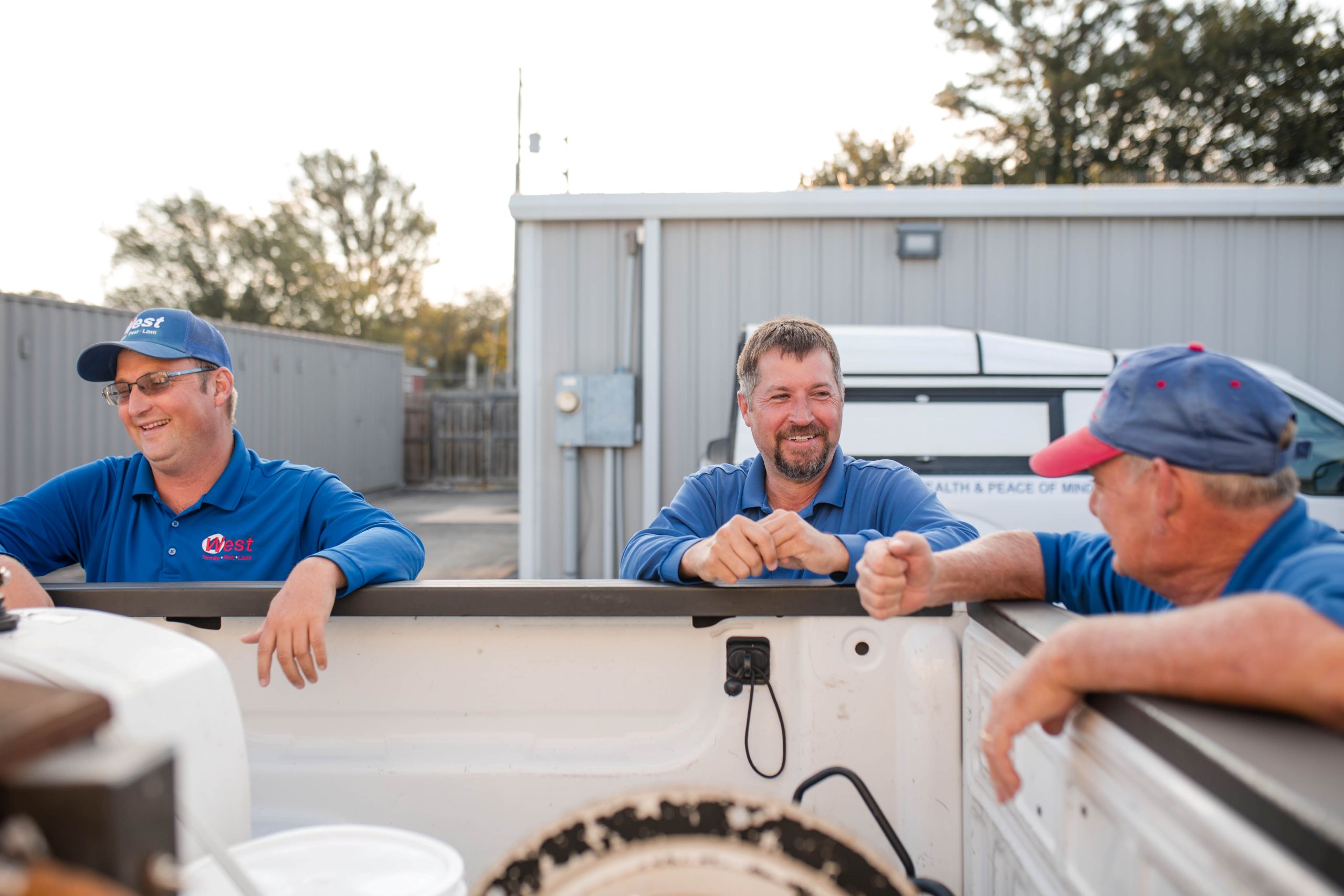 Three men in blue shirts and hats lean on the side of a white pickup truck, smiling and talking about pest solutions in a parking lot with a building and van in the background.