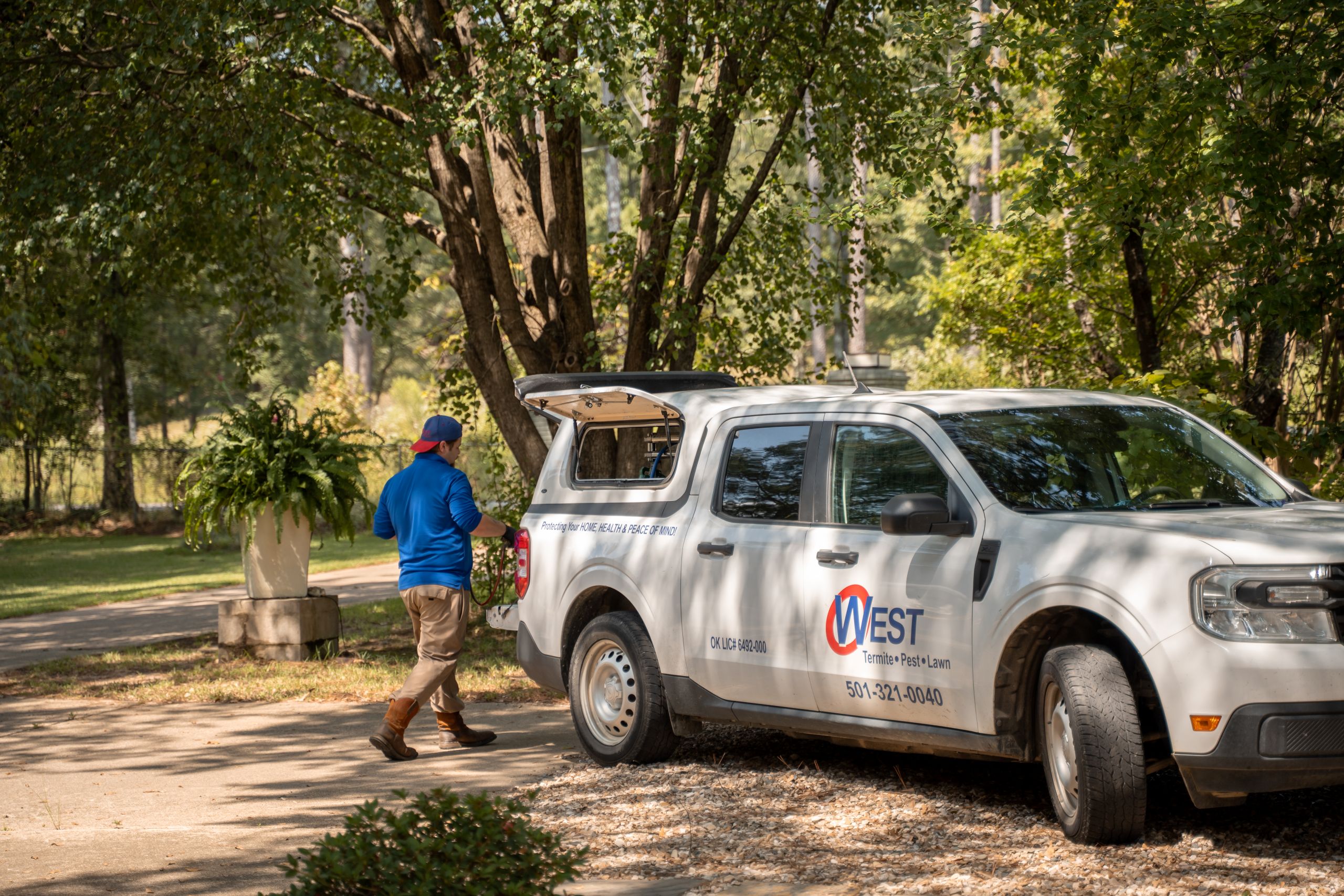 Why Lawn Care Matters Before Cooler Weather Arrives A person in boots and a blue shirt walks toward a white West Termite, Pest & Lawn truck parked on a shaded driveway near trees and plants.