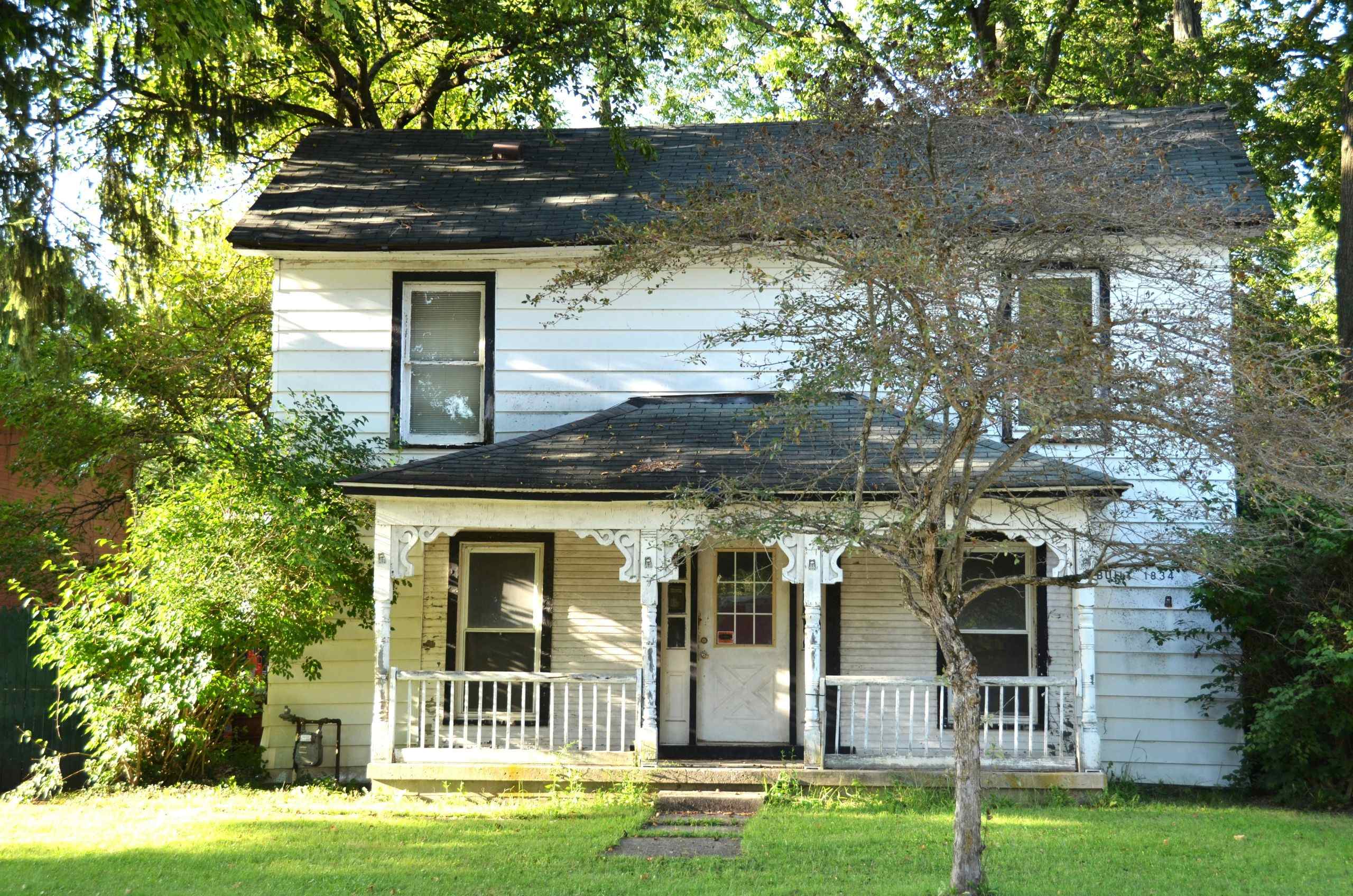 A two-story white house with a covered front porch and gingerbread trim, featuring a date plaque that reads Built 1834.