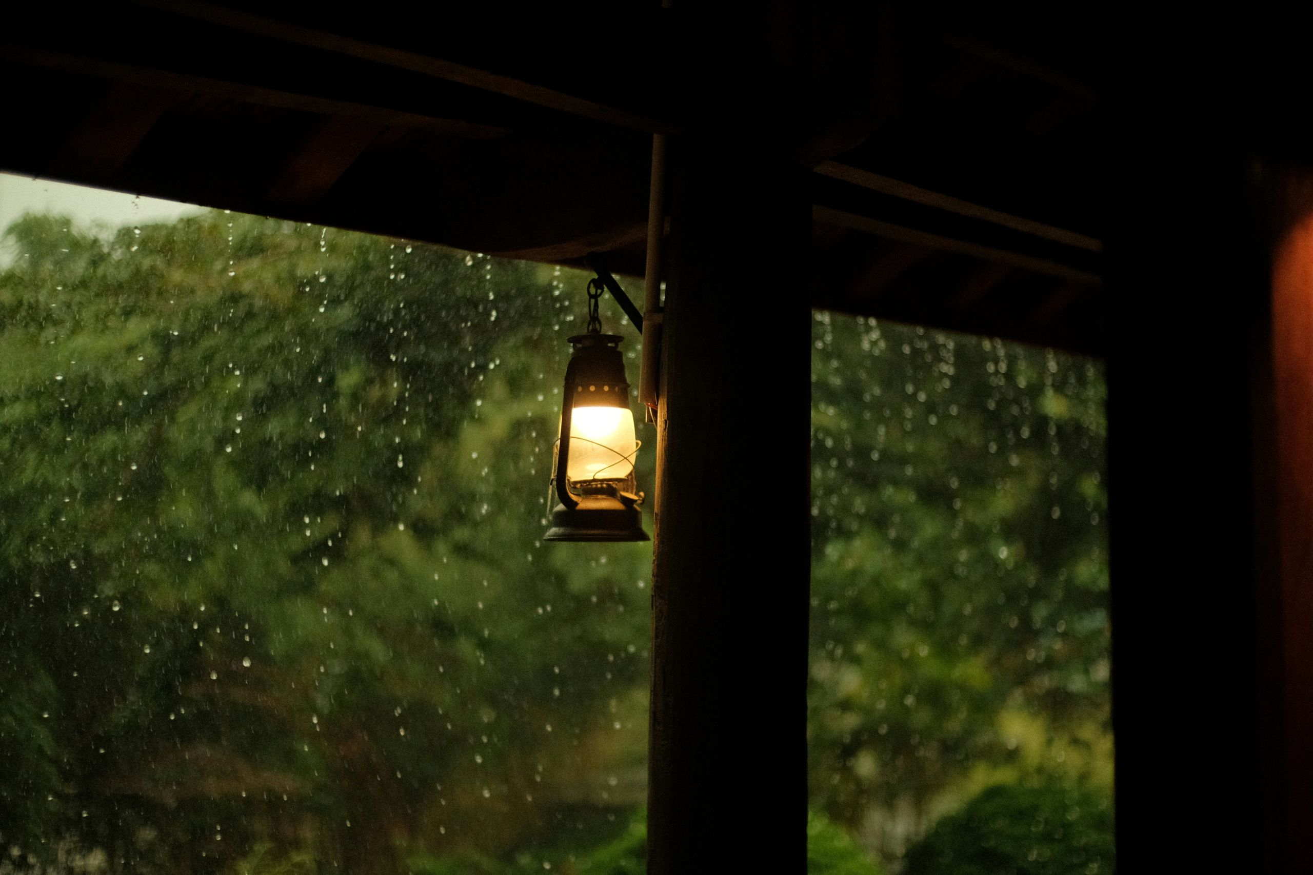 A glowing lantern hangs from a post on a porch during a rainstorm.